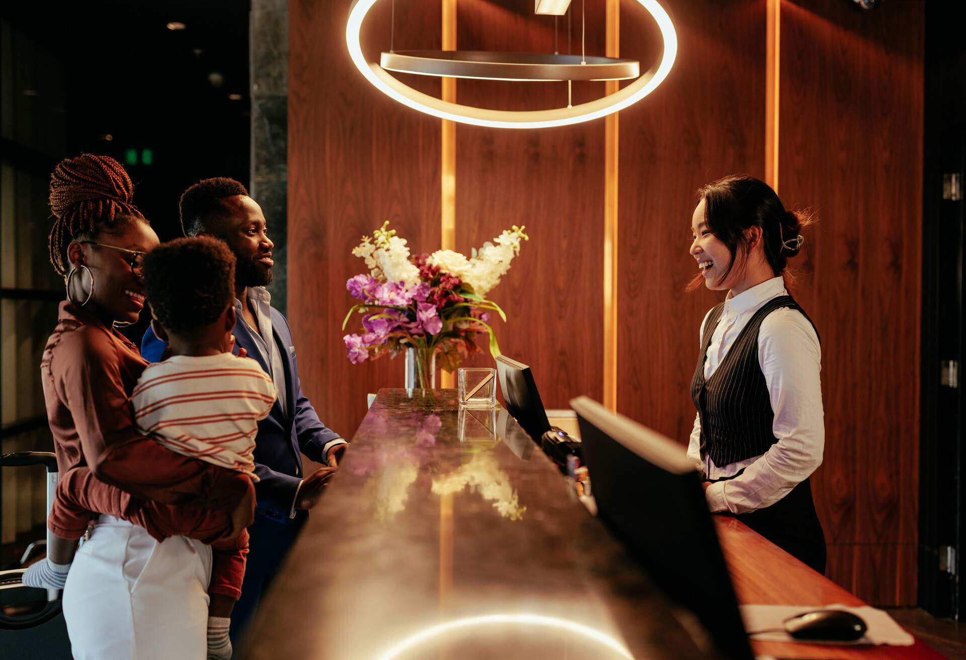 A happy couple with their young son stands at a hotel reception desk while a hotel clerk welcomes and assists them with the check-in process.