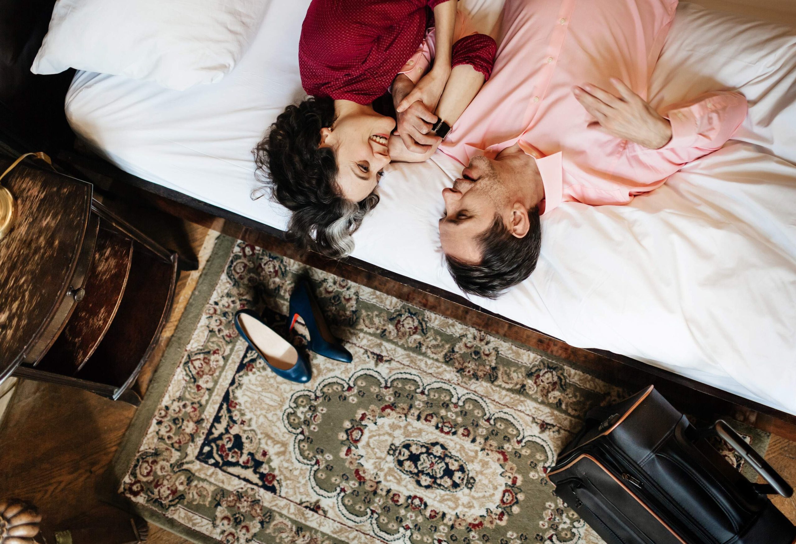 A mature, loving couple is looking at each other lying on the white sheets of a hotel bed. They are holding hands and seem to be in love. On the floor, there's an old carpet, a suitcase and the woman's shoes.