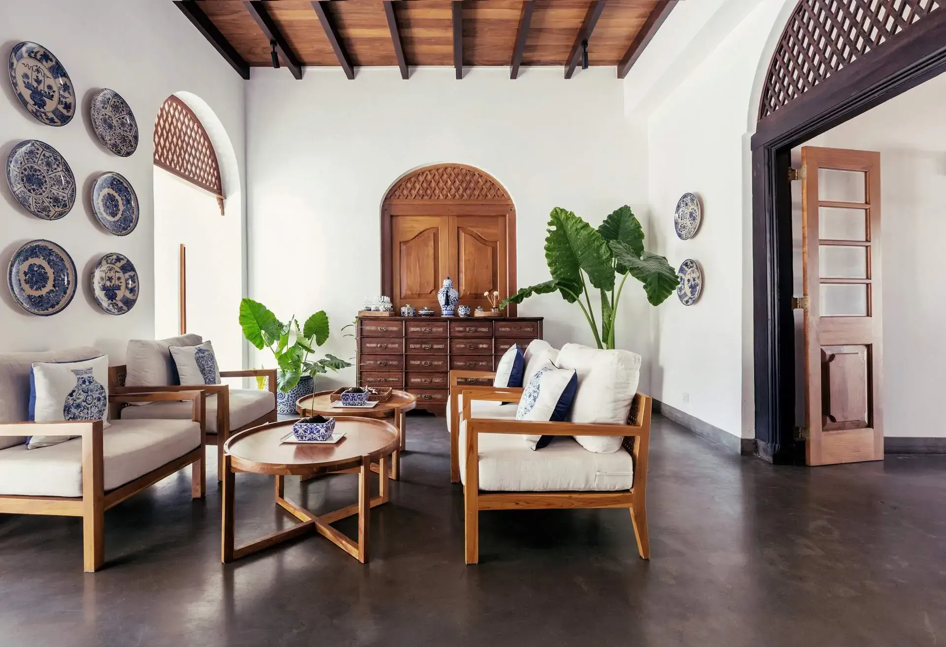 A light and bright living area with seats, wooden coffee tables, potted plants, and decorative wall plates.